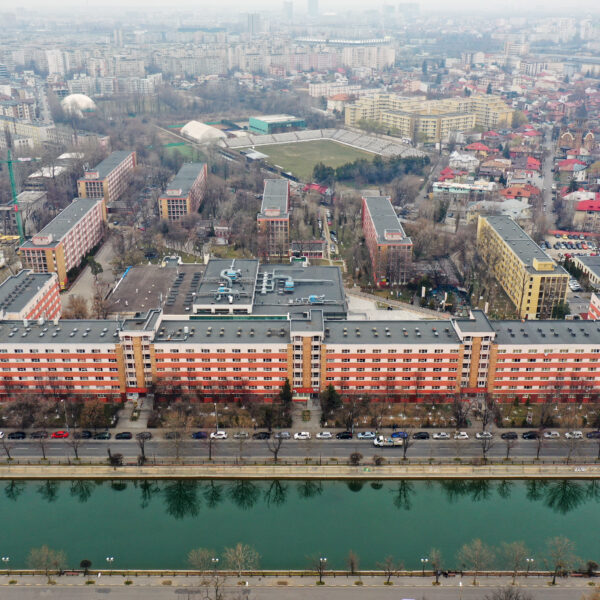 Aerial view of a city with a wide river in the foreground, mid‑rise buildings, and a large red brick apartment block along the road.