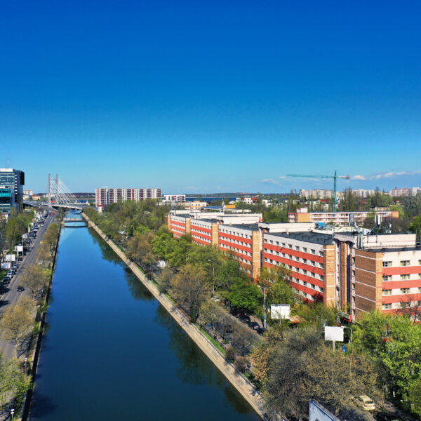 Aerial cityscape with a long canal flanked by buildings and trees under a bright blue sky.