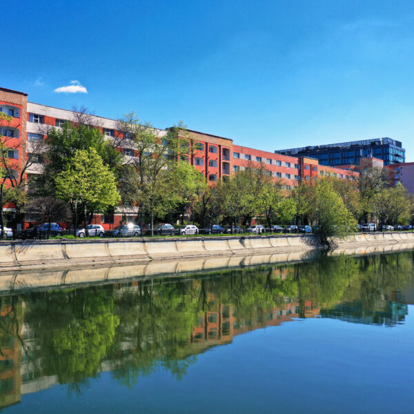 Row of red-brick apartment buildings beside a calm canal, with green trees and a bright blue sky; modern glass towers in the distance.
