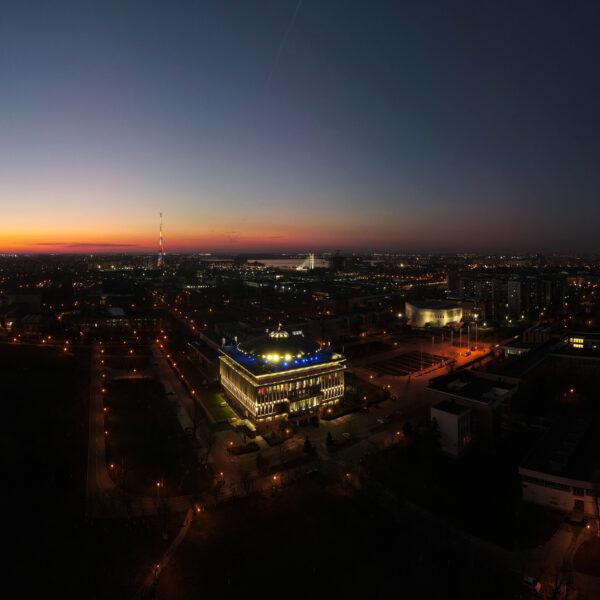 Panoramic city skyline at dusk with a gradient orange to dark blue sky and illuminated streets and buildings in the foreground.