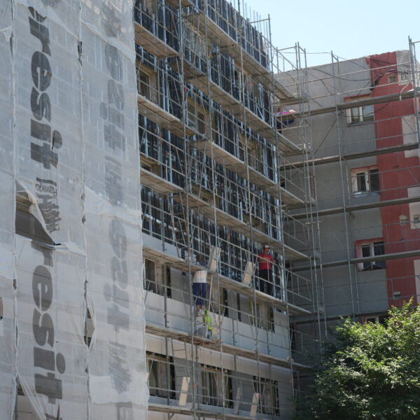Construction site: a multi-story building under renovation with scaffolding and white protective wrap, workers in hard hats visible on the façade.