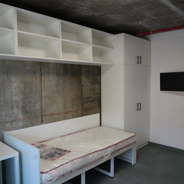 White modular storage with open cubbies and tall cabinets along a concrete wall, a wrapped mattress on a white bed frame in front, in a modern showroom.