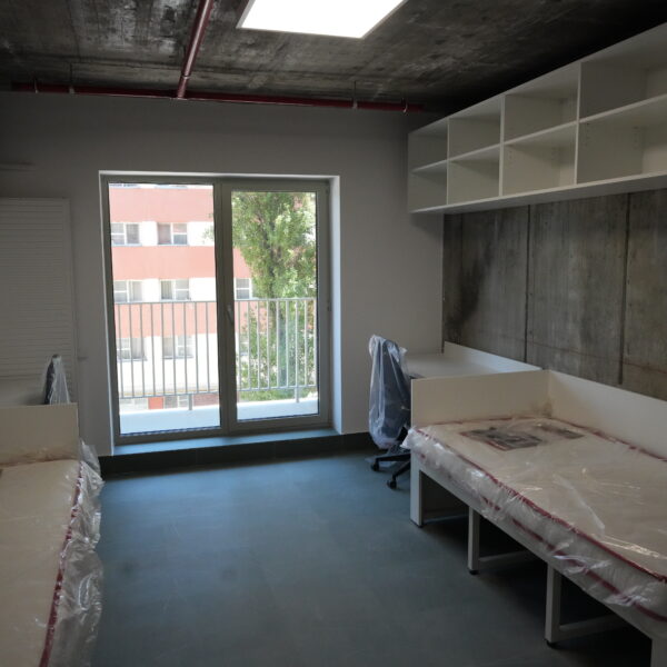 Empty room under construction with white open shelving along the wall, concrete accent wall, and desks wrapped in plastic near a balcony window.
