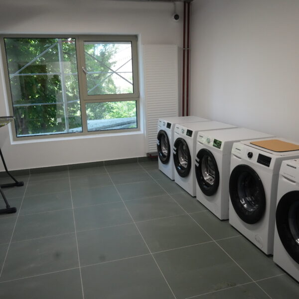 Laundry room with four front-loading washing machines lined up along the right wall and ironing boards on the left, near a large window showing greenery outside.