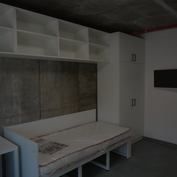 Interior showroom with white cabinets and open shelves above a concrete wall; bed frames with plastic-wrapped mattresses and a wall-mounted TV nearby.
