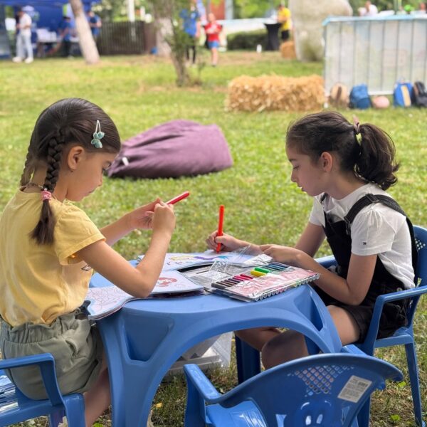 Two girls coloring at a small blue table outside, with markers and coloring sheets in a grassy park near festival decorations.