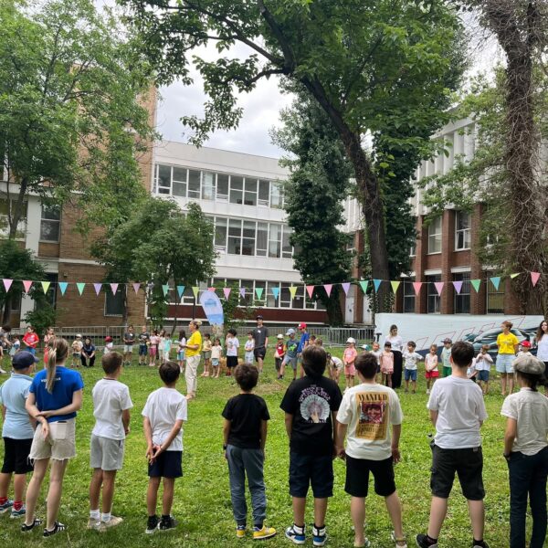 Group of children standing in a circle on a grassy school yard, colorful bunting overhead and brick buildings in the background.