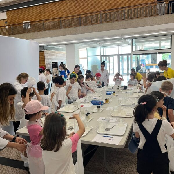 Group of children in white lab coats working at long tables with laboratory equipment, supervised by adults in a bright indoor space.