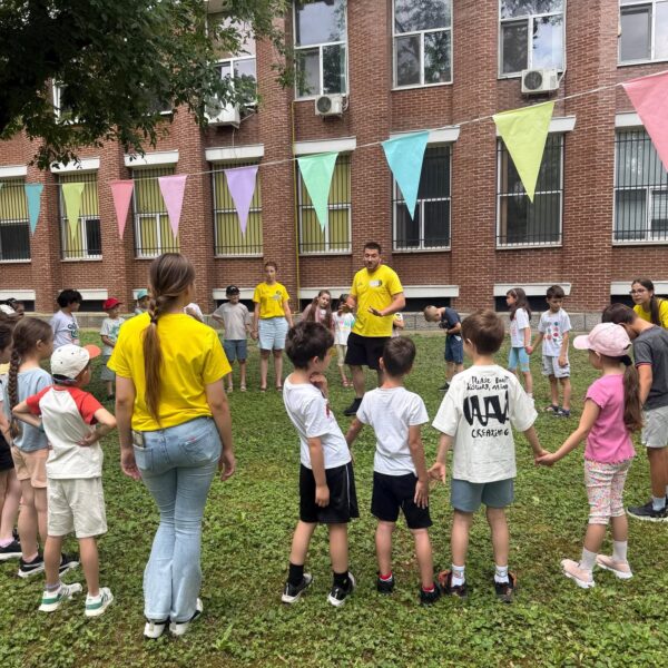 Children stand in a circle on a grassy area outside a brick building, with pastel pennant flags strung overhead.