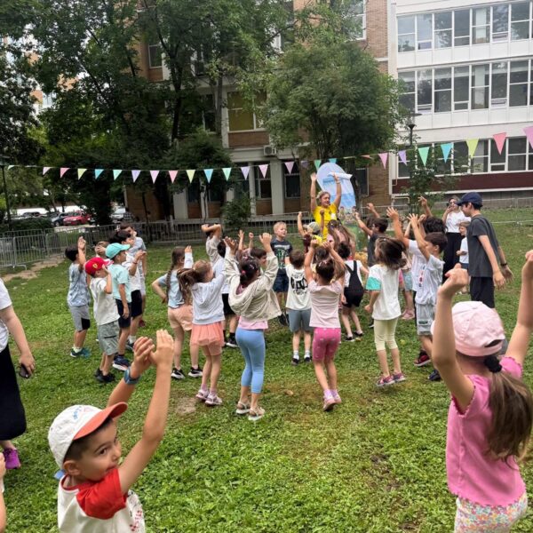 Group of children standing in a circle on a grassy schoolyard, hands raised as a teacher leads an outdoor activity, colorful bunting strung overhead and a building in the background.